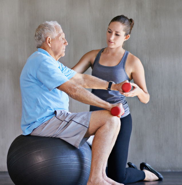 Trainerin betreut einen Senior beim Hanteltraining auf dem Gymnastikball
